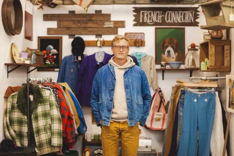 Mężczyzna w sklepie z odzieżą vintage, gdzie wisi szyld "FRENCH CONNECTION". W tle wiszą ubrania i dekoracje.