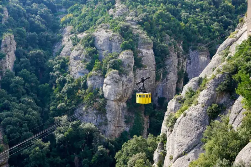 A bright yellow cable car ascends the rugged, tree-covered cliffs of Montserrat, Spain.
