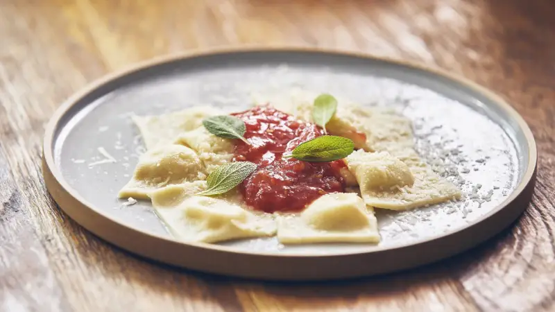 Pyszne raviolini z sosem pomidorowym i parmezanem, ozdobione listkami szałwii, na szarym talerzu.