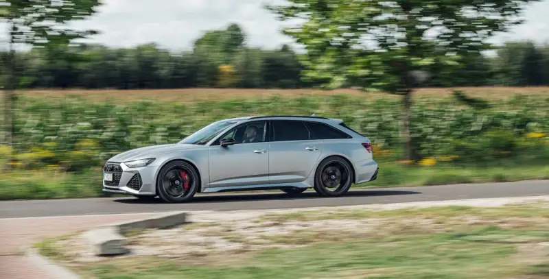 Srebrne Audi RS6 Avant pędzi po wiejskiej drodze, otoczone zielenią pól i drzew.