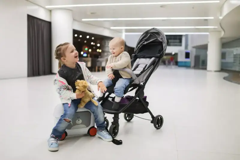 Dzieci w podróży z Ryanair. Starsza siostra siedzi na walizce z misiem, młodsza w wózku.