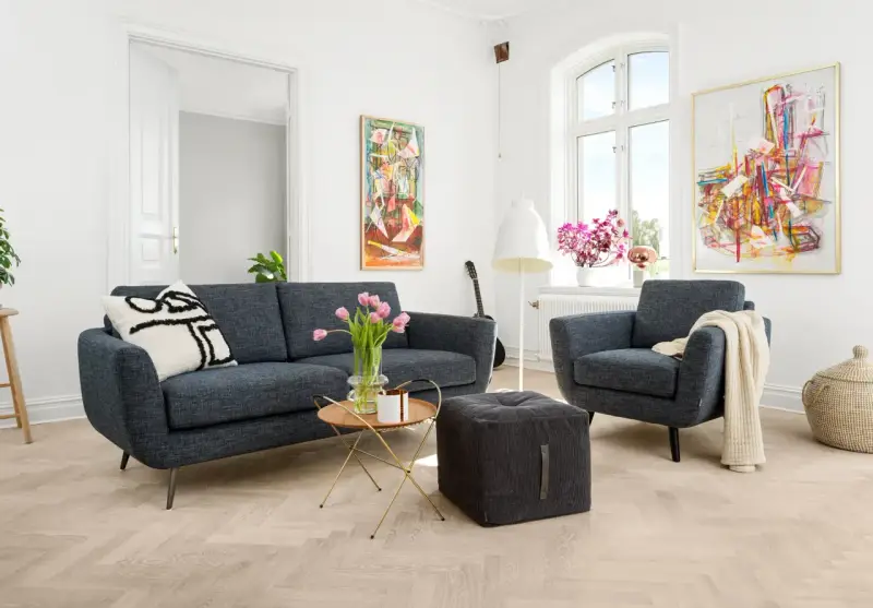 En modern soffgrupp 3+2 i mörkblått tyg, med en fåtölj, soffbord, pall och konst på väggarna.