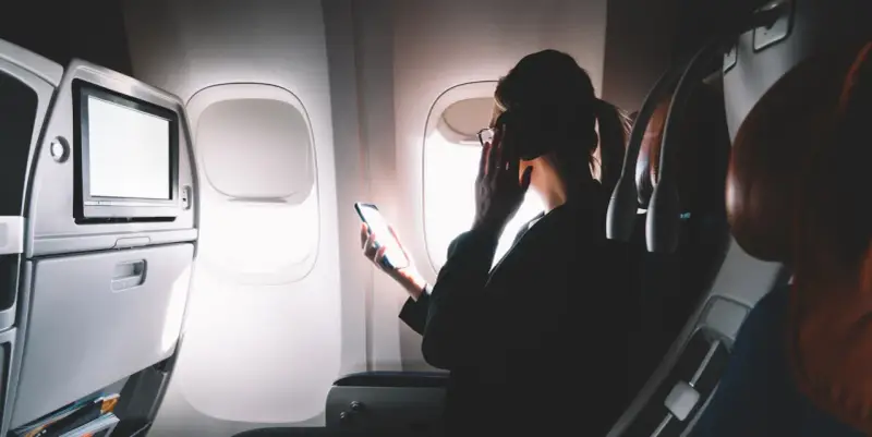 Kobieta w okularach trzyma telefon, zastanawiając się, czy w samolocie jest wifi. Obok ekran rozrywki.