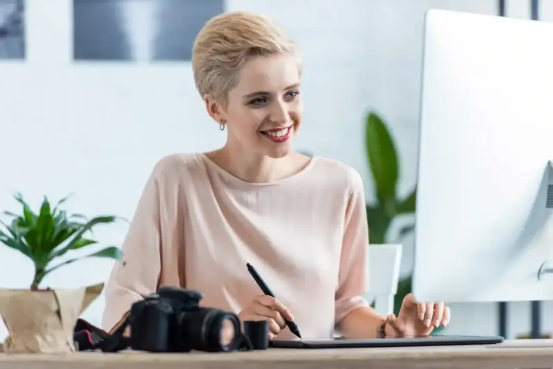 Uśmiechnięta kobieta z krótkimi blond włosami pracuje przy komputerze, obok aparat fotograficzny. Sprawdza najnowsze oferty pracy w Pile.
