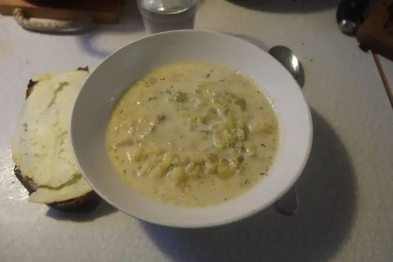 En krämig fänkålssoppa med potatis serveras i en vit skål, bredvid en bit rostat bröd med ost.