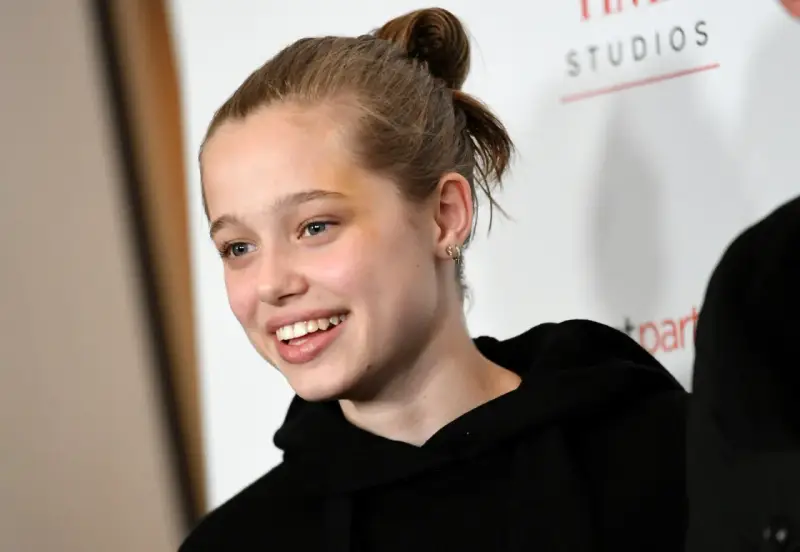Uśmiechnięta Shiloh Jolie Pitt z włosami upiętymi w kok, w czarnej bluzie z kapturem.
