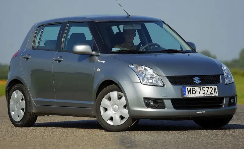 Szary Suzuki Swift 2007 z kierowcą za kierownicą, zaparkowany na drodze.