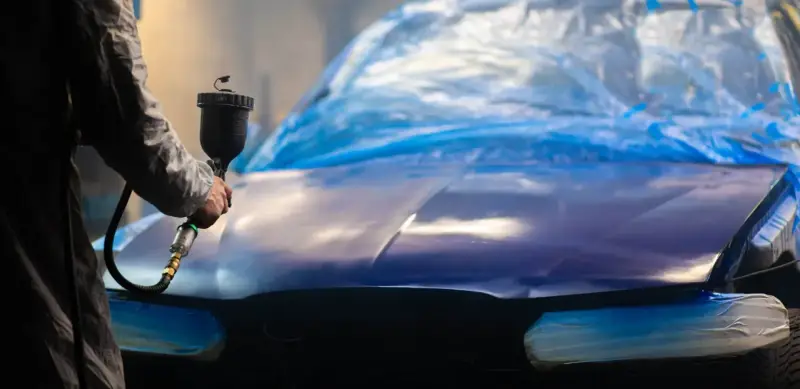 Person lackar om bilen billigt med sprutpistol. Bilen är täckt med blå plast.