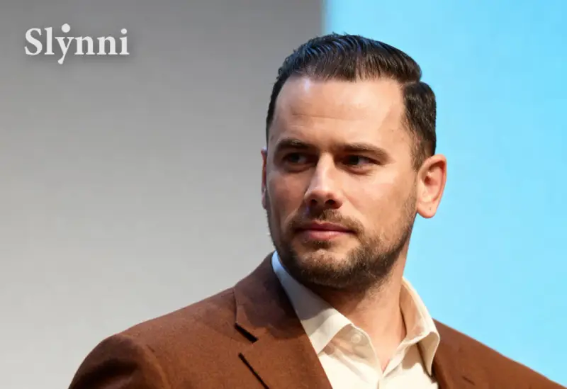 Grzegorz Collins, znany z programów telewizyjnych, w eleganckim brązowym garniturze. Jego wiek jest trudny do określenia, ale prezentuje się dojrzale.