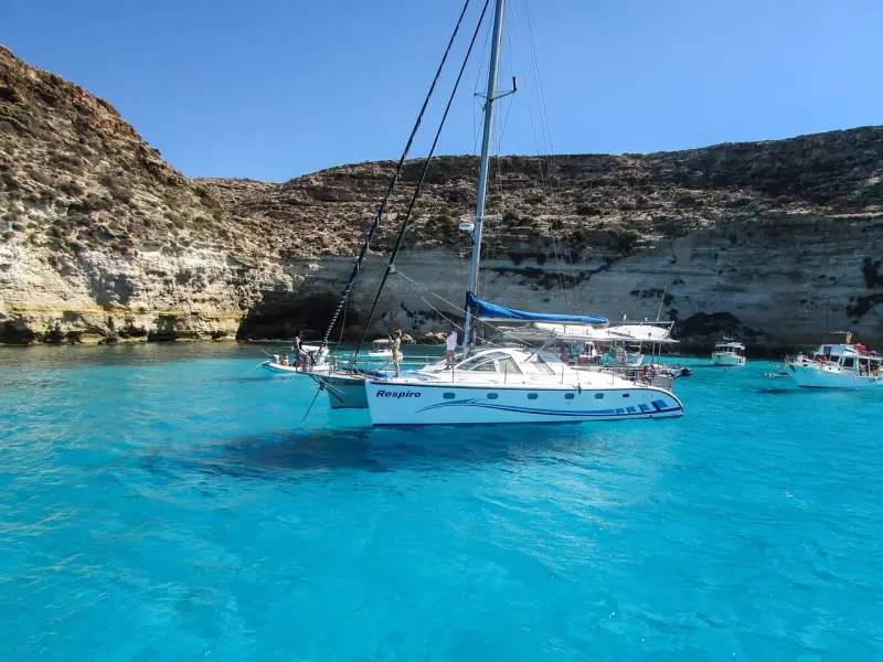Catamarano "Respiro" in acque cristalline di un'isola sotto la Sicilia, con scogliere rocciose e altre barche.