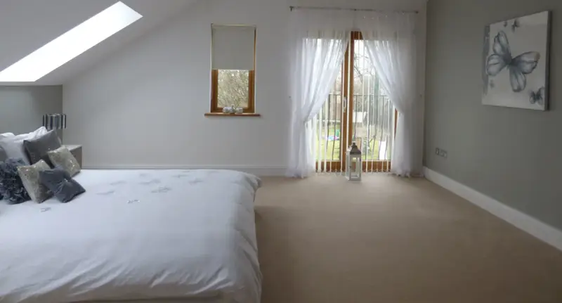 Dormitorio con cama blanca, ventana abuhardillada y puerta acristalada con cortinas. La orientación oeste de los pisos permite una luz suave.
