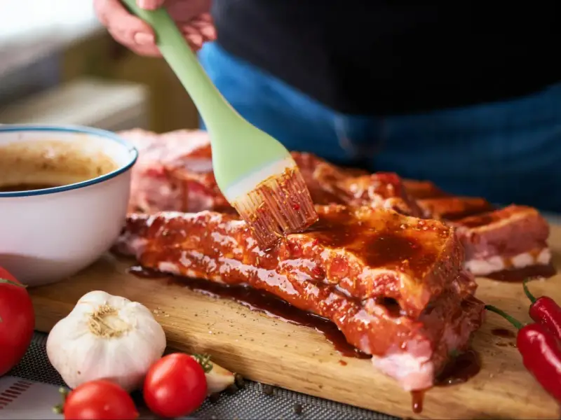 Pędzel rozprowadza aromatyczną marynatę do żeberek na surowym mięsie. Obok czosnek, pomidorki i papryczki chili.