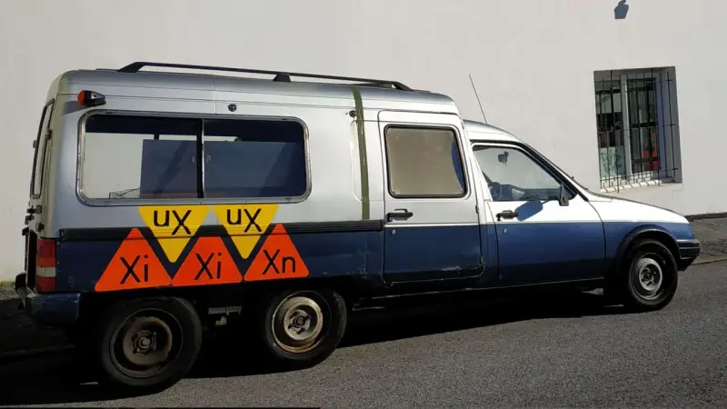 Dwukolorowy Citroen C15 z nietypowymi oznaczeniami "UX" i "Xi Xn" na boku, zaparkowany przy białej ścianie.