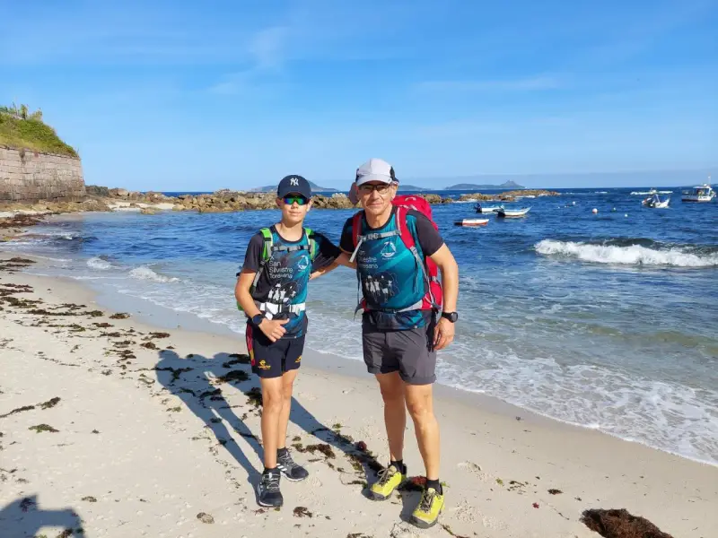 Dos excursionistas, padre e hijo, disfrutan de la playa en la etapa Baiona Vigo. El mar azul y el cielo despejado acompañan su aventura.
