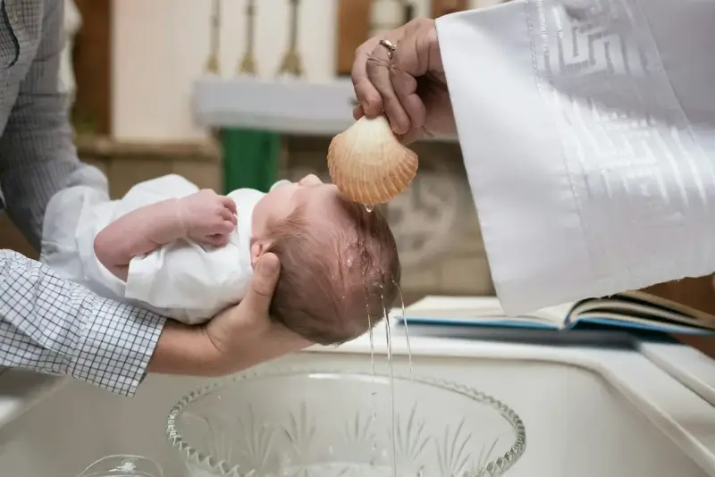 Chrzestna polewa główkę dziecka wodą święconą z muszli podczas ceremonii chrztu.