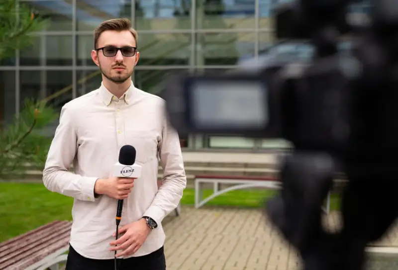 Młody dziennikarz w okularach trzyma mikrofon z logo "FLESZ" przed kamerą.