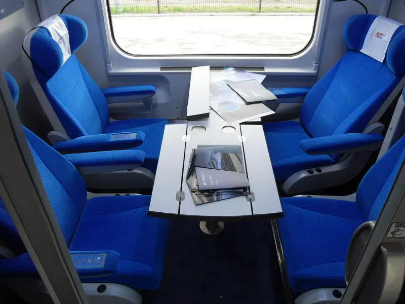 Wnętrze wagonu przedziałowego z wygodnymi, niebieskimi fotelami i stolikiem.