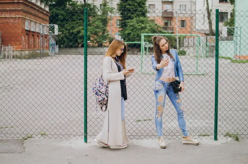 Dwie dziewczyny w modnych stylizacjach, idealnych na outfit do szkoły, stoją przy płocie. Jedna robi sobie selfie, druga przegląda telefon.