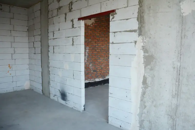 Wnętrze w budowie. Widoczny otwór drzwiowy z czerwonym nadprożem stalowym, ściany z białych pustaków i cegły.