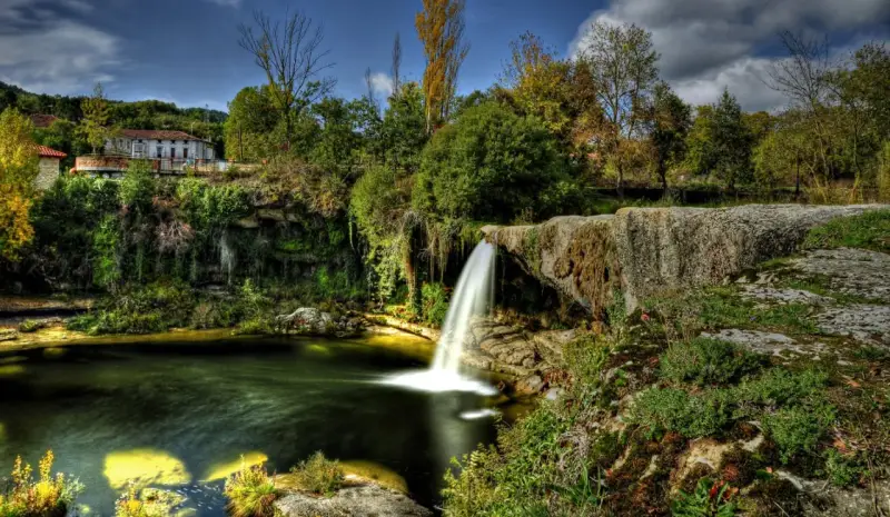 Cascadas en España: una casa rural se asoma entre la vegetación exuberante junto a una cascada que cae en un estanque de aguas verdes.