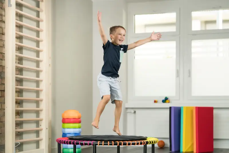 Fröhlicher Junge springt auf Trampolin, Teil der kinder physiotherapie. Kletterwand und bunte Matten im Hintergrund.