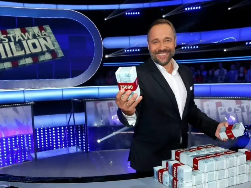Uśmiechnięty prowadzący teleturniej trzyma w dłoni paczkę pieniędzy. Na stole piętrzą się stosy banknotów.