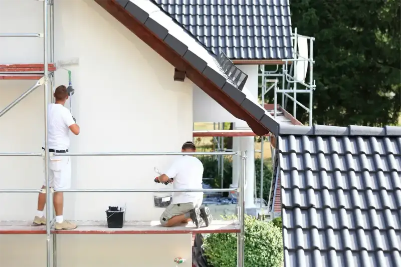 Dwóch mężczyzn na rusztowaniu maluje białą elewację domu. Sprawdź, jaka jest malowanie elewacji cena.