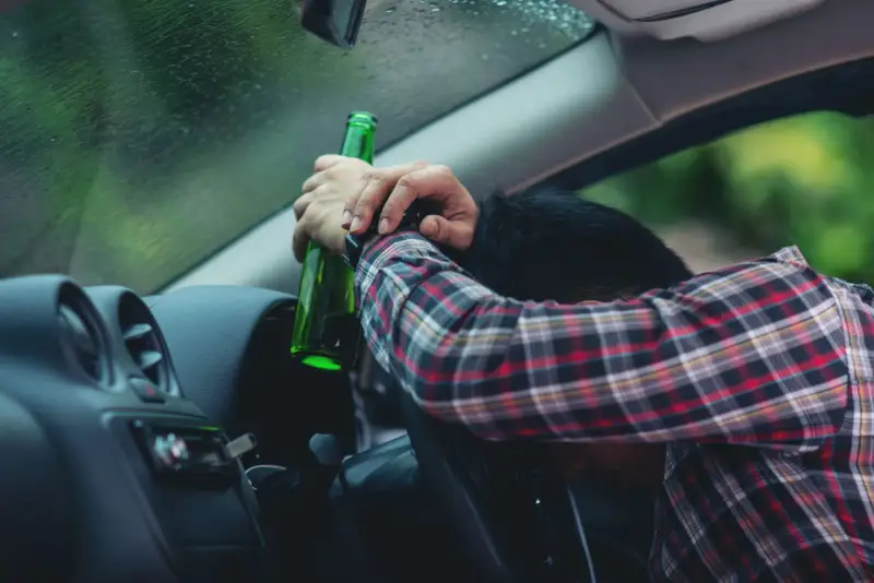 Mężczyzna w kratę trzyma butelkę, opierając głowę na kierownicy. To obraz ostrzegawczy przed jazdą pod wpływem alkoholu, przypominający o zagrożeniach i potrzebie blokady alkoholowej.
