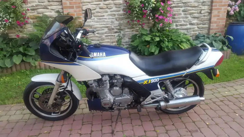 Klasyczny motocykl Yamaha XJ 900 w kolorze białym i granatowym, z niebieskimi i żółtymi pasami, zaparkowany na brukowanym podjeździe.