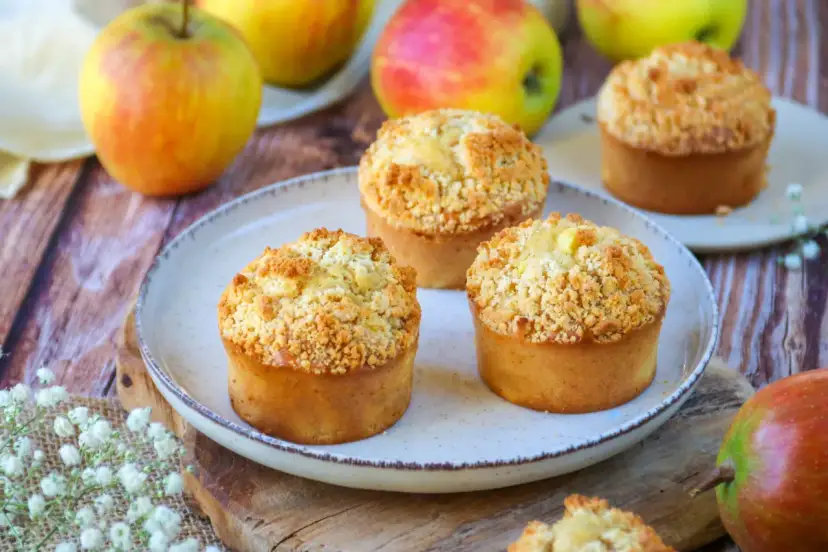 Trois muffins pomme amande dorés avec un crumble croustillant, entourés de pommes fraîches sur une table en bois.