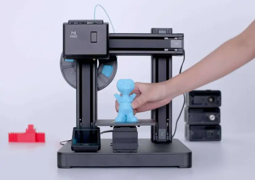 Drukarka 3D z niebieską figurką i ręką wyjmującą ją z platformy.