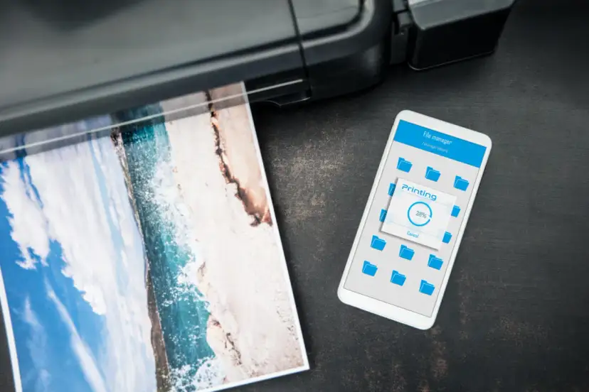Jak wydrukowac cos z iPhone? Widok telefonu z ekranem "Printing 38%" obok drukarki drukującej zdjęcie plaży.