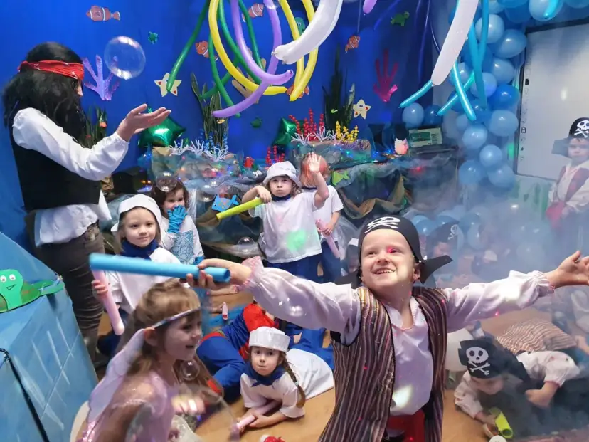 Dzieci w strojach piratów i wróżek bawią się na podwodnym balu karnawałowym. Balony i dekoracje tworzą atmosferę morskiej przygody.