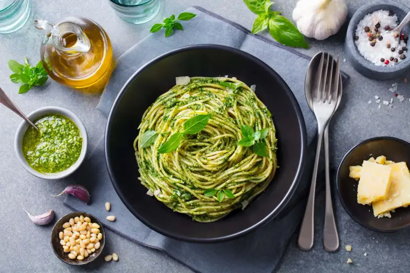Spaghetti z pesto i bazylią w czarnej misce, obok sos, orzeszki piniowe, czosnek i parmezan.