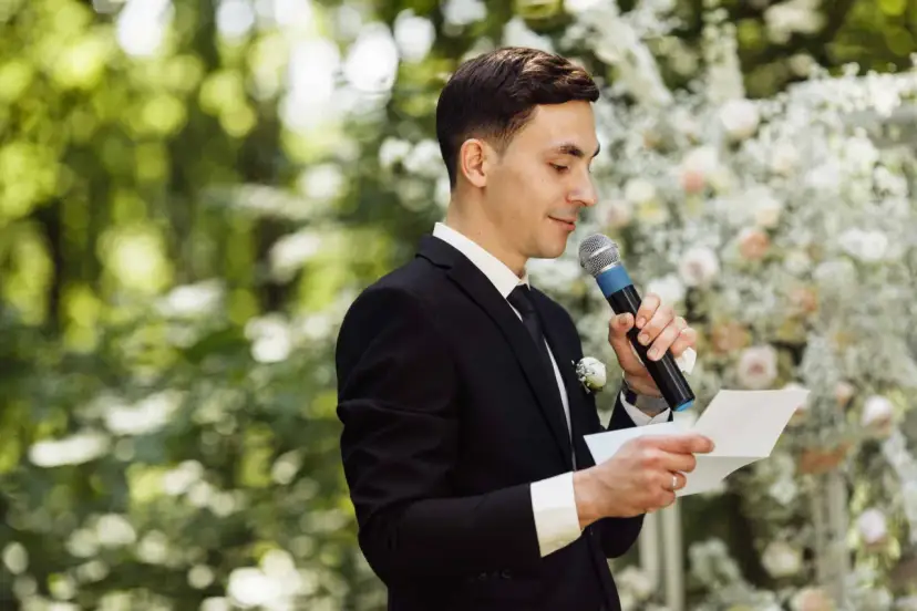 El maestro de ceremonias, con traje negro y micrófono, lee un discurso frente a un arco de flores.