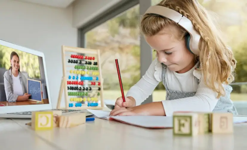 Dziewczynka w słuchawkach uczy się matematyki online, pisząc w zeszycie. Na ekranie laptopa nauczycielka pokazuje zadanie.