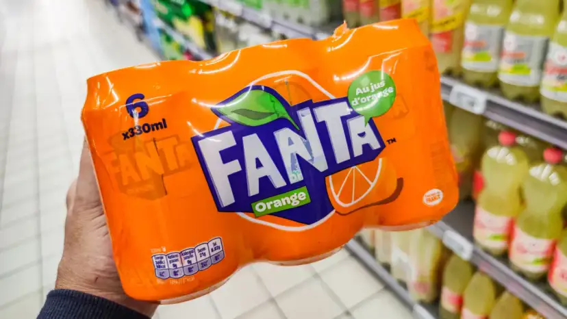 Opakowanie 6 puszek Fanty Orange w ręce, na tle sklepowych półek z napojami.