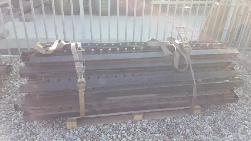 Stos czarnych, metalowych elementów naczepy z pasami transportowymi.