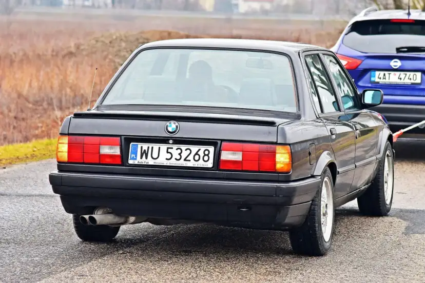 Tył czarnego BMW E30 z tablicą rejestracyjną WU 53268, holowanego przez niebieskiego SUV-a.