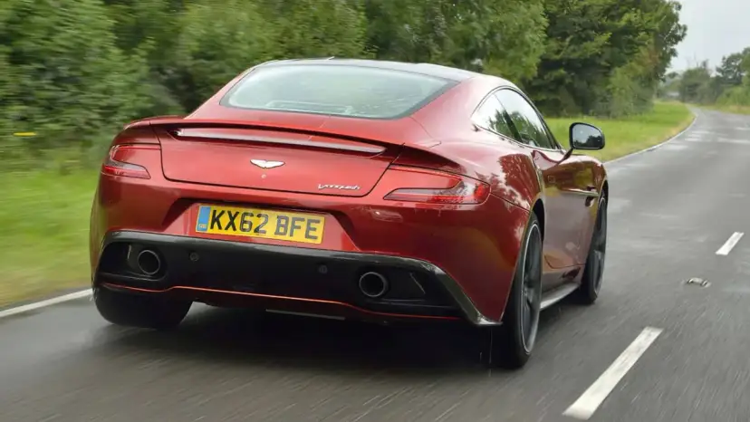 Czerwony Aston Martin Vanquish pędzi po mokrej drodze, otoczony zielenią.