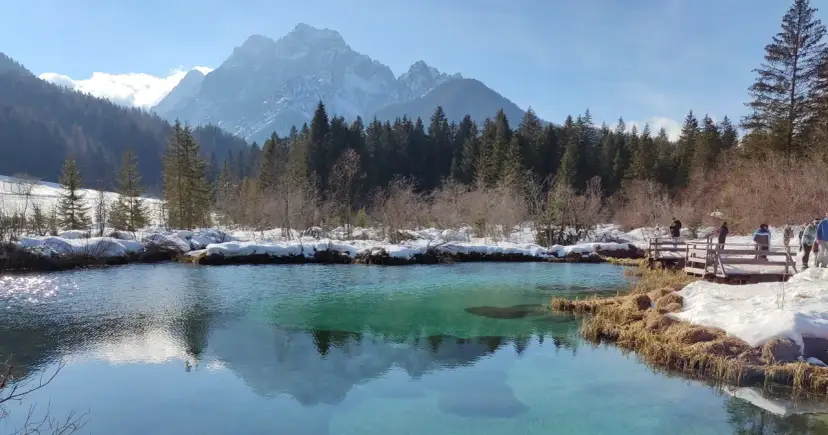 Turkusowa woda i ośnieżone szczyty gór zdradzają, gdzie leży Planica. Widok na zimowy krajobraz z drewnianą kładką i turystami.