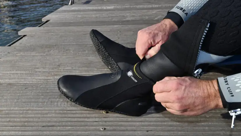 Mężczyzna zakłada czarne buty neoprenowe na drewnianym pomoście przy wodzie.