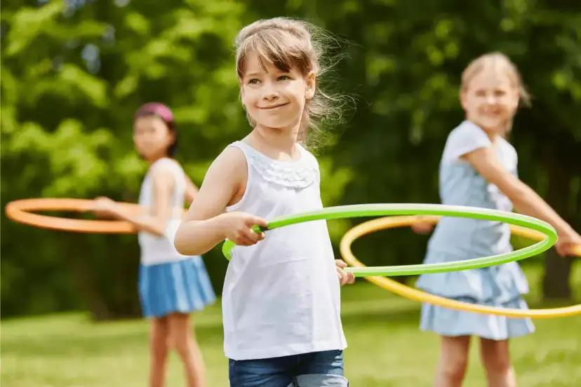 Uśmiechnięte dziewczynki biorą udział w konkurencjach sportowych dla dzieci, kręcąc hula-hop na trawie.