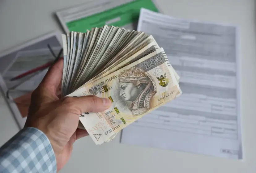 Dłoń trzyma plik banknotów 200 zł. W tle dokumenty, być może dotyczące tego, ile godzin to 0.5 etatu.