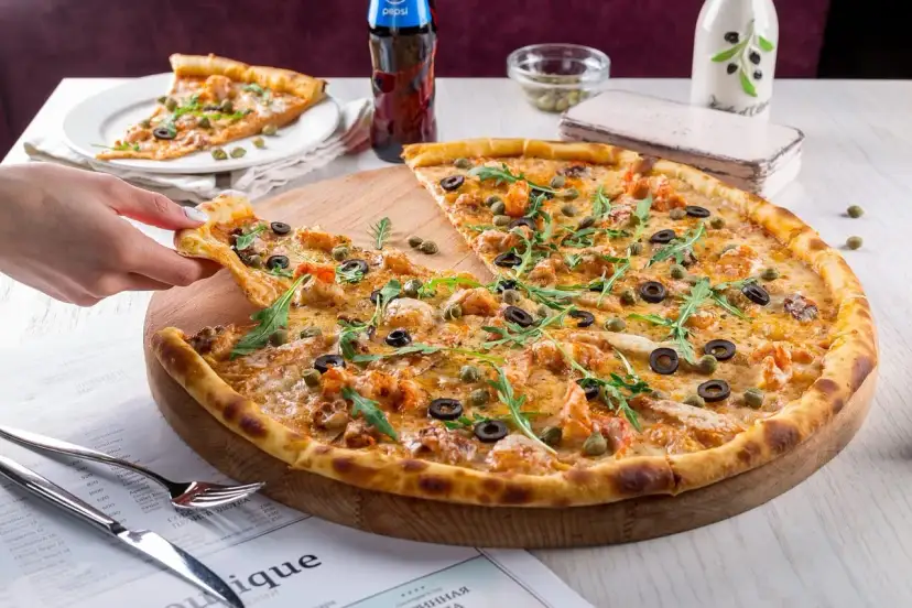 Pizza z krewetkami, oliwkami i kaparami na drewnianej desce, ręka podnosi kawałek.