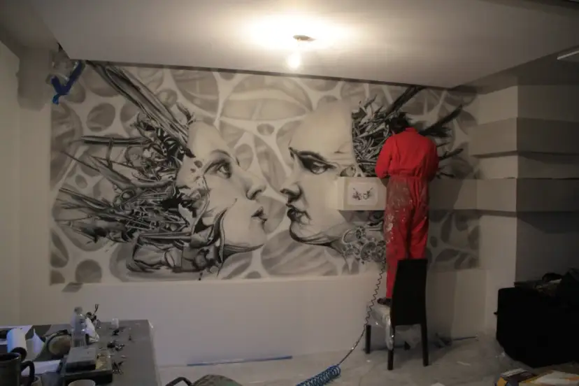 Artysta w czerwonym kombinezonie tworzy malarstwo artystyczne wnętrz – surrealistyczny mural z twarzami i mechanicznymi elementami.