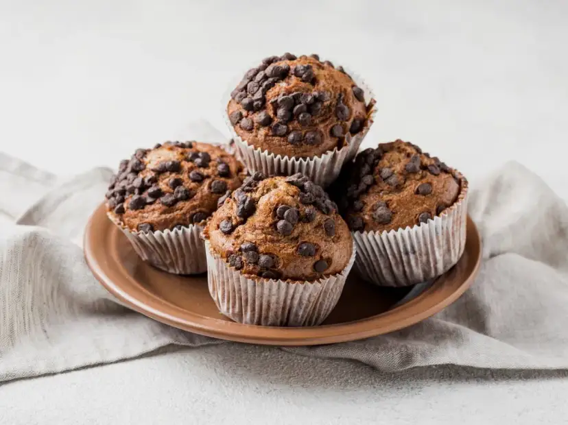 Cztery muffinki z kawałkami czekolady na brązowym talerzu, na szarym obrusie.