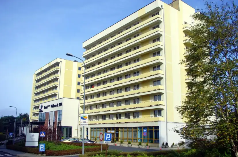 Hotel Vestina w Międzyzdrojach, wysoki, żółty budynek z balkonami, otoczony zielenią.