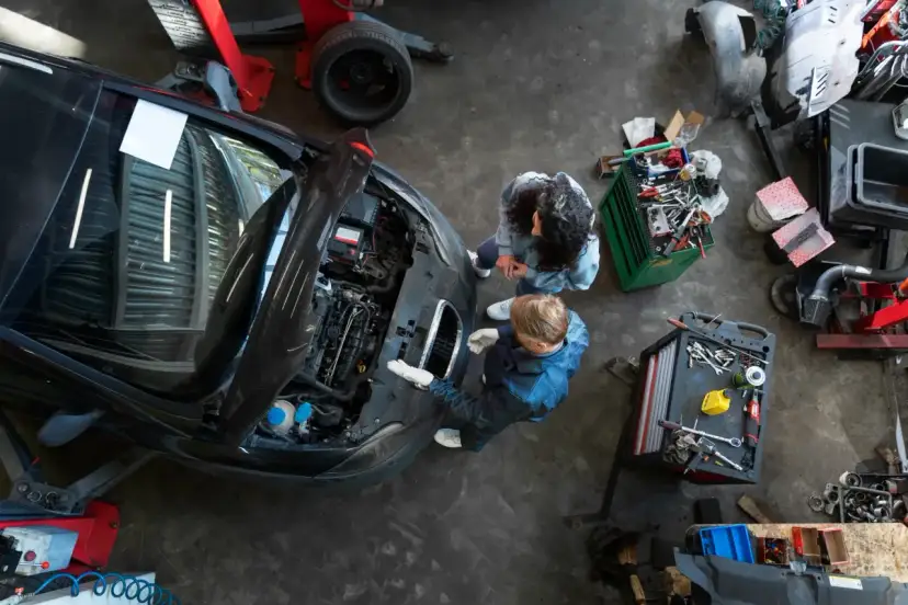 Warsztat samochodowy z dwoma mechanikami przy otwartej masce czarnego auta i narzędziami.