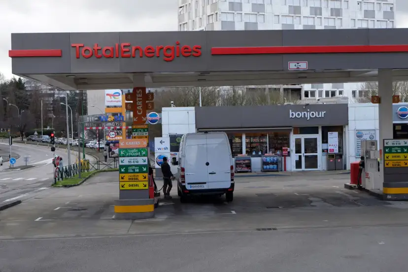 Station TotalEnergies avec une camionnette blanche en cours de ravitaillement. Panneaux de prix et de services.
